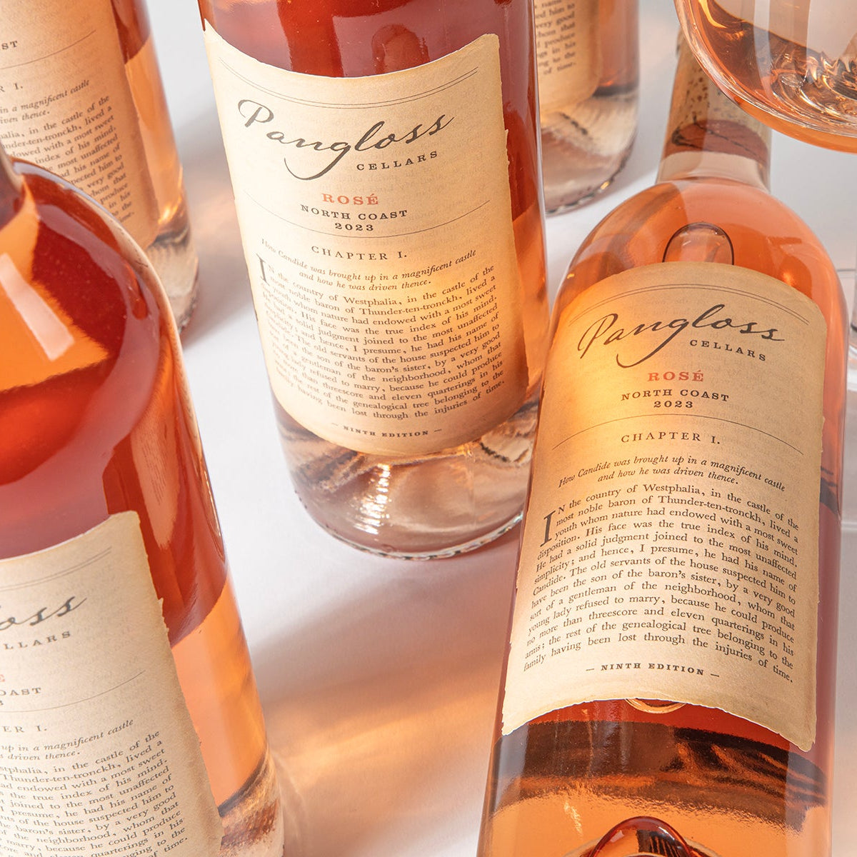 Several bottles of Paragloss Cellars rosé wine with cream labels and a glass of rosé wine on a white surface, photographed in warm, clean studio lighting.