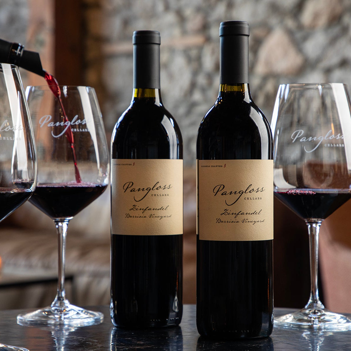 Two bottles of Pangloss wine with glasses being poured and filled, set against a blurred background.