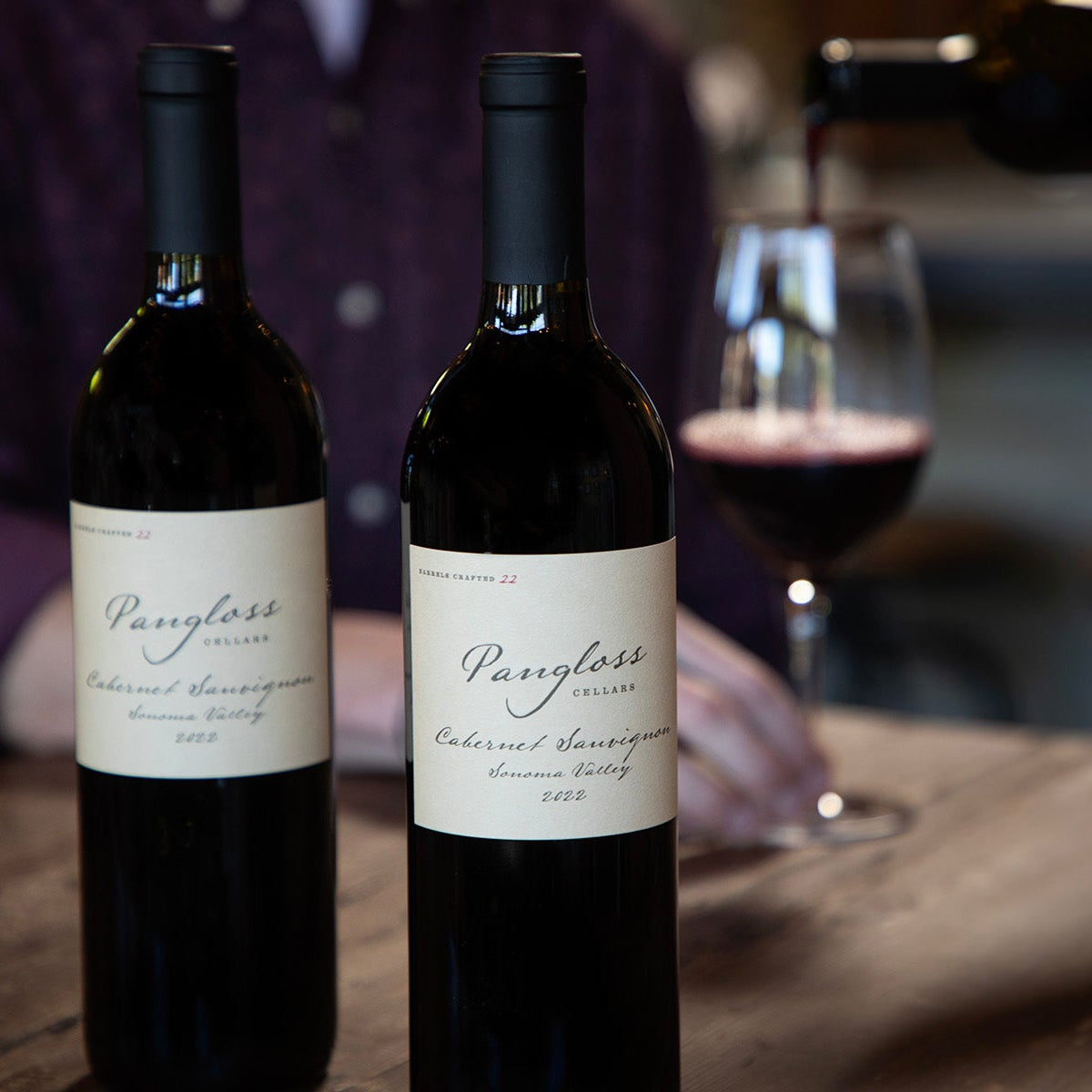 Two bottles of Pangloss Cabernet Sauvignon wine on a wooden table with a blurred glass of wine in the background.