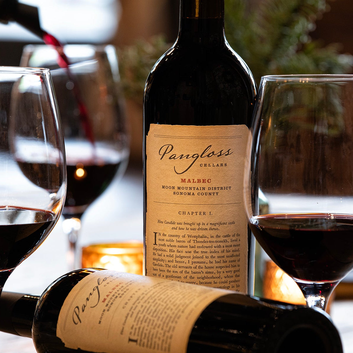 Two bottles and two glasses of red wine with a third bottle being poured, on a wooden table.