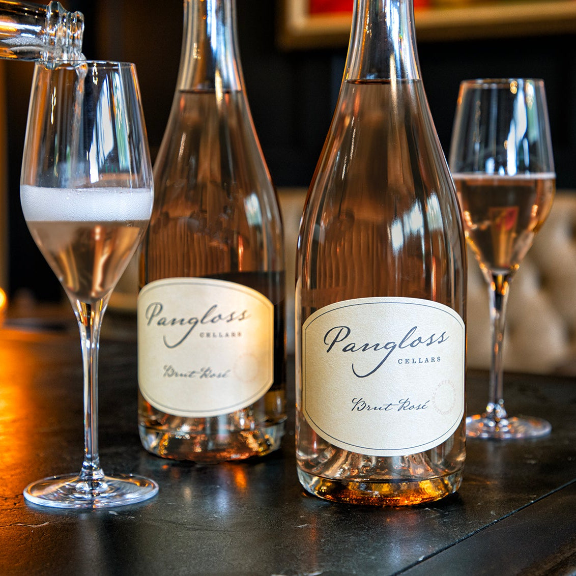 Two bottles of Pangloss sparkling rosé wine with glasses on a bar counter.