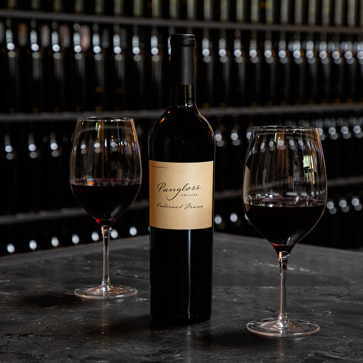 Wine bottle with two glasses on a dark surface in front of wine racks.