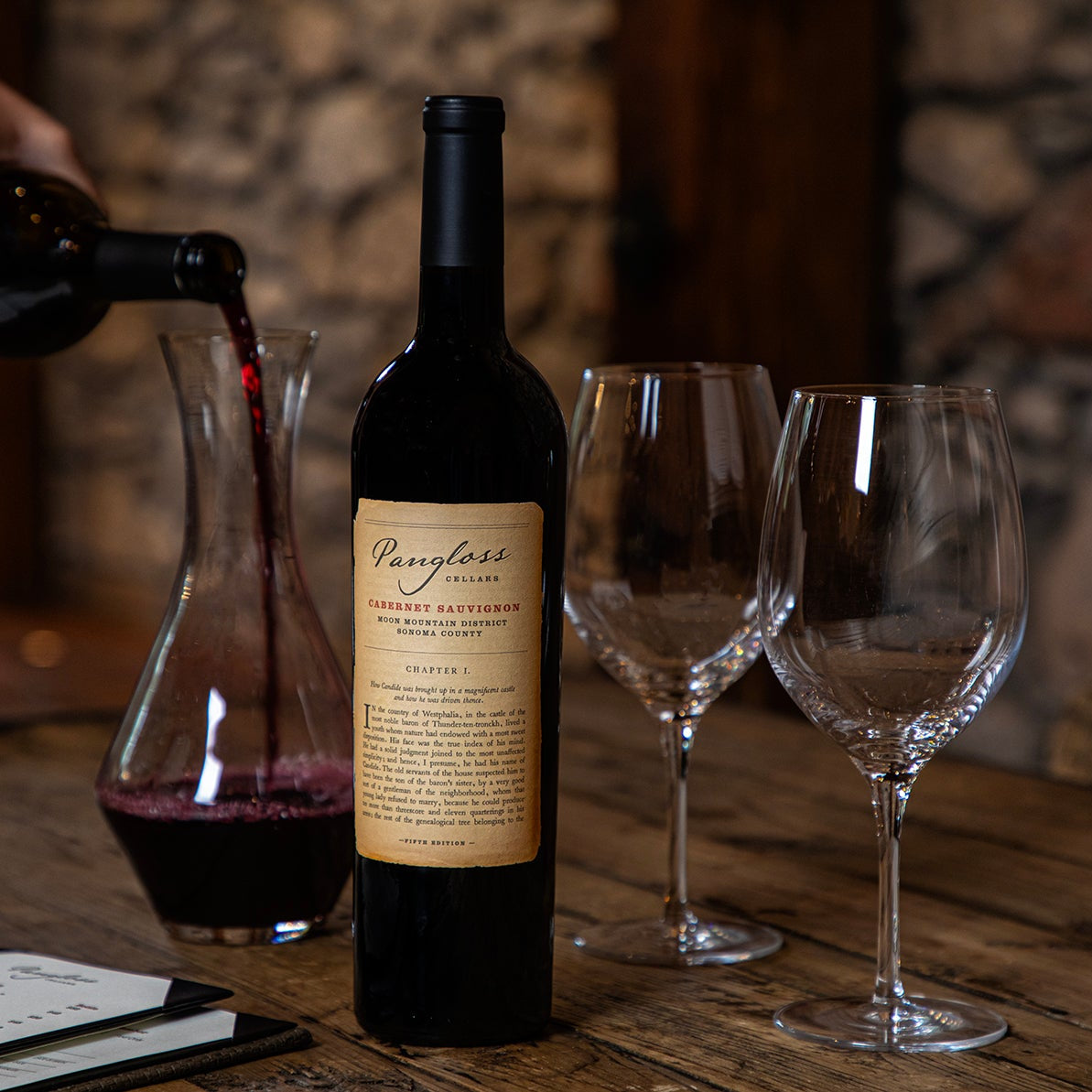 Person pouring red wine into a glass with a bottle of Pangloss wine on a wooden table.