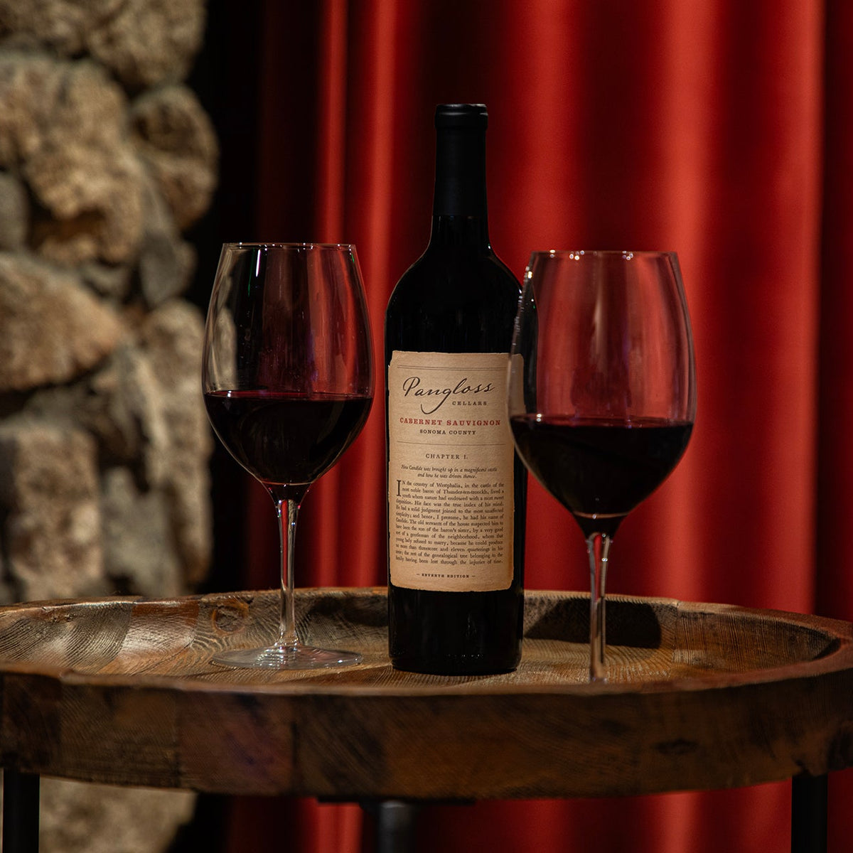 Wine bottle and two glasses of red wine on a wooden table with a stone wall and red curtain background.