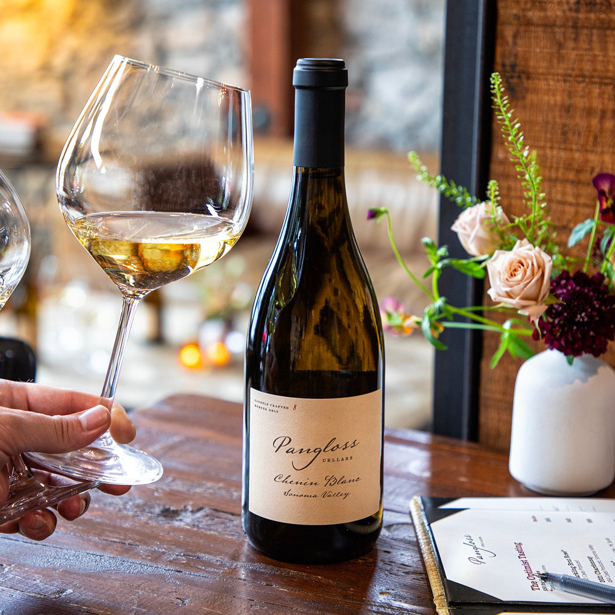 Wine bottle and glasses on a table with a hand holding a glass, flowers in the background
