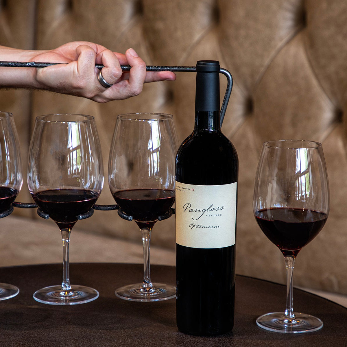 Person opening a bottle of wine with four glasses filled with red wine on a table.