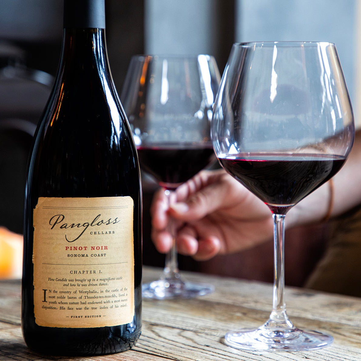 Pangloss wine bottle with two glasses of red wine on a wooden table.
