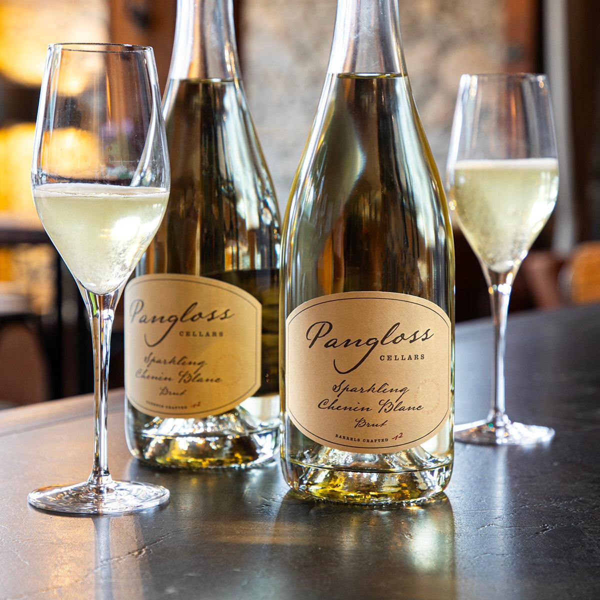 Two bottles of Pangloss Cellars sparkling wine with two glasses on a bar.