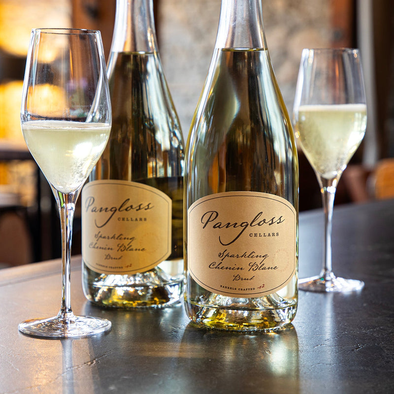 Two bottles of Pangloss Cellars sparkling wine with two glasses on a bar.