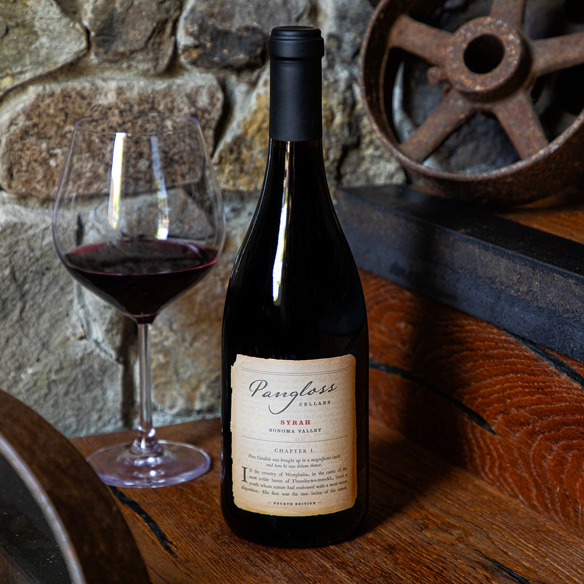 Bottle of red wine with a glass on a wooden surface against a stone wall.