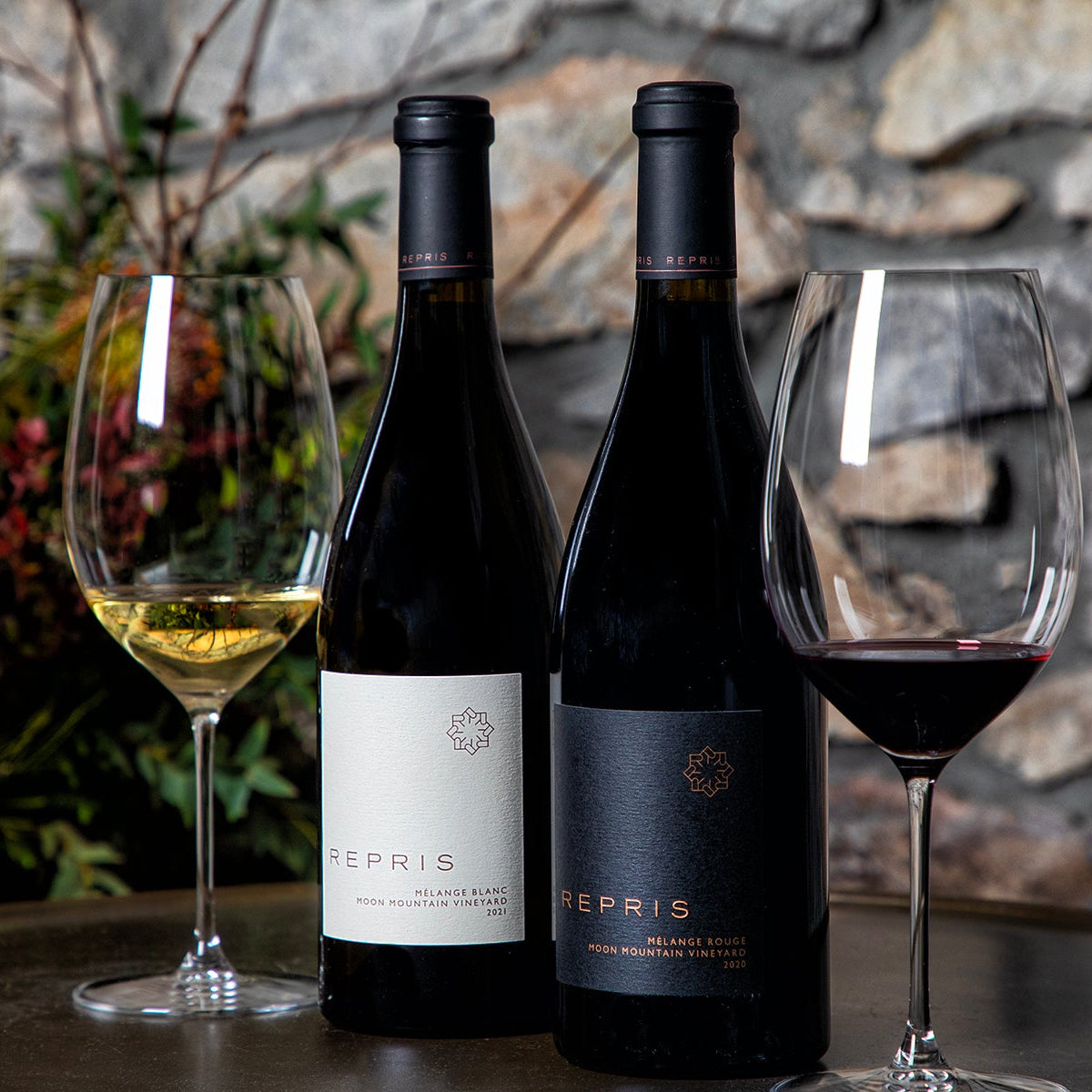 Two bottles of Repris wine, one white and one red, displayed on a table with two wine glasses in front of a rustic stone wall background.