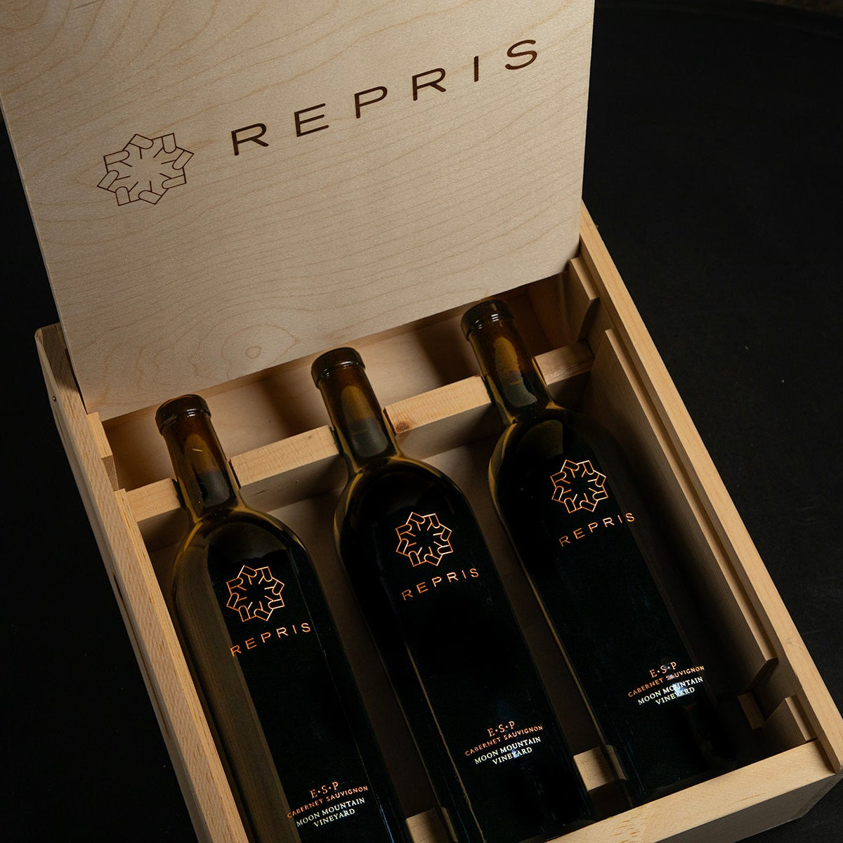 Three wine bottles in a wooden box with 'Repris' branding on a dark background