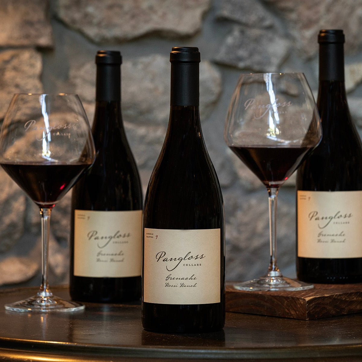 Two bottles of wine with labels and two glasses filled with red wine on a stone surface.