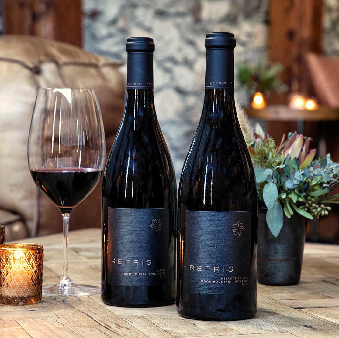 Two dark wine bottles labeled 'REPRIS' sit on a wooden table beside a filled wine glass, lit candles, and a small floral arrangement in a cozy indoor setting with leather seating in the background.