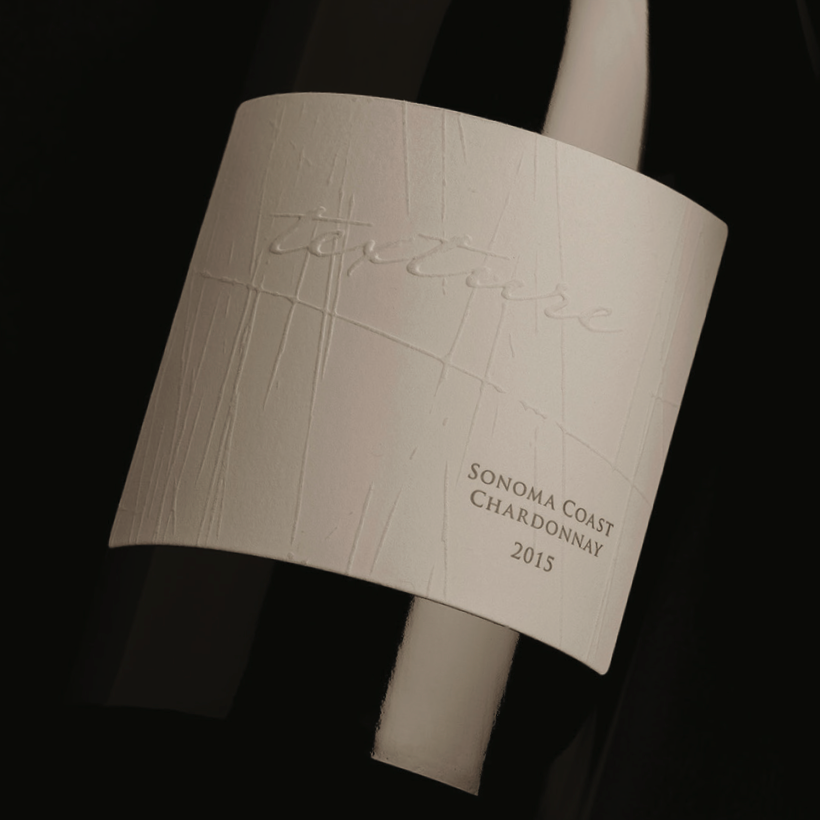 A close-up of a textured off-white wine label wrapped around a dark bottle, with embossed script and printed text reading 'Texture Sonoma Coast Chardonnay 2015'.